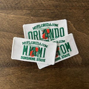 May include: Three white embroidered patches with the text "MYFLORIDA.COM" and the names of Florida cities: "ORLANDO", "MIAMI", and "TAMPA". Each patch features a green palm tree with an orange sun and the text "Sunshine State".