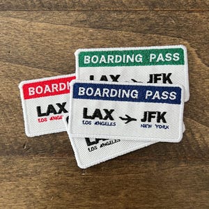 May include: Four white fabric patches with blue, red, and green borders. The patches are stacked on top of each other. The top patch says "Boarding Pass" in green letters. The second patch says "Boarding Pass" in blue letters. The third patch says "LAX JFK" in black letters. The bottom patch says "LAX JFK" in black letters with "Los Angeles" and "New York" written below.