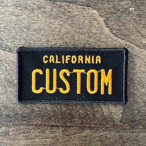 May include: Black fabric patch with yellow embroidered text that reads "CALIFORNIA CUSTOM".