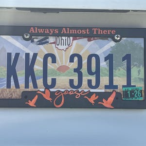 May include: Black license plate frame with the text "Always Almost There" and a sunset scene with the text "Ohio" and the license plate number "KKC 3911". The frame also has a goose silhouette with the word "goose" in cursive.