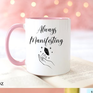 May include: White ceramic mug with a pink rim and handle. The mug has a black design of a hand holding a crystal with the text "Always Manifesting".