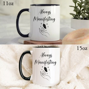 May include: White ceramic mug with a black rim and handle. The mug has a black design of a hand holding a crystal with stars around it. The text "Always Manifesting" is printed in black on the mug. The mug is available in two sizes: 11 oz and 15 oz.