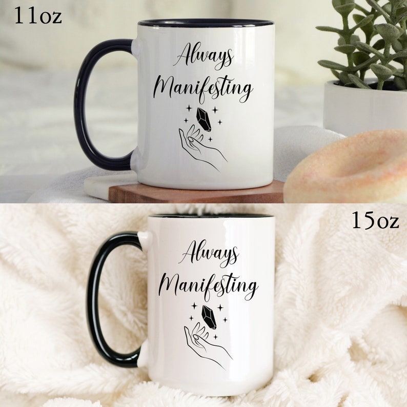 May include: White ceramic mug with a black rim and handle. The mug has a black design of a hand holding a crystal with stars around it. The text "Always Manifesting" is printed in black on the mug. The mug is available in two sizes: 11 oz and 15 oz.