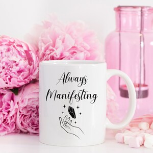 May include: White ceramic mug with black text that reads "Always Manifesting" and a black illustration of a hand holding a crystal with stars. The mug is surrounded by pink peonies and pink marshmallows.