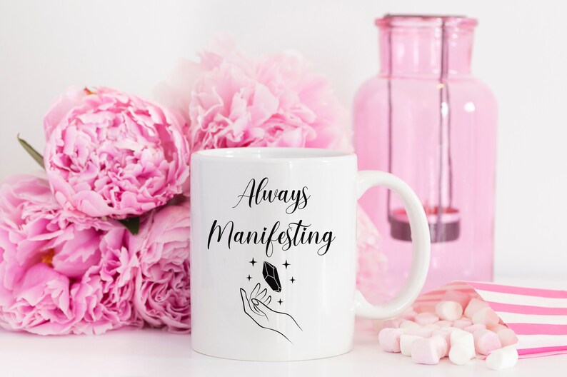 May include: White ceramic mug with black text that reads "Always Manifesting" and a black illustration of a hand holding a crystal with stars. The mug is surrounded by pink peonies and pink marshmallows.