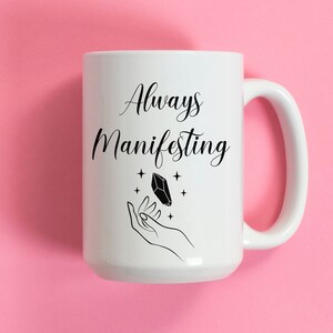 May include: White ceramic coffee mug with black text that reads "Always Manifesting" and a black illustration of a hand holding a crystal.