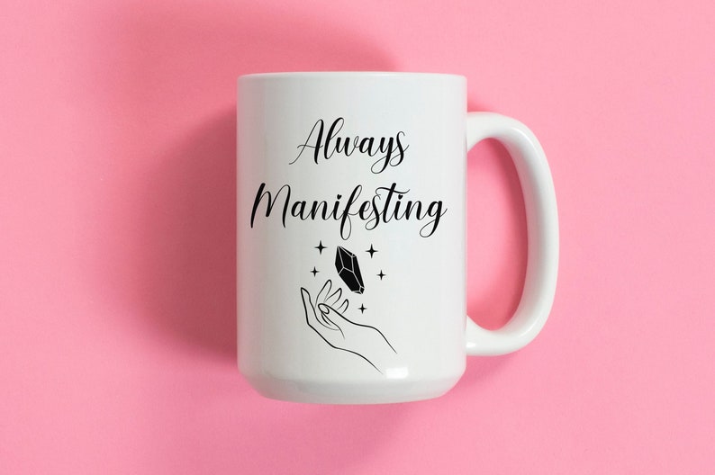 May include: White ceramic coffee mug with black text that reads "Always Manifesting" and a black illustration of a hand holding a crystal.