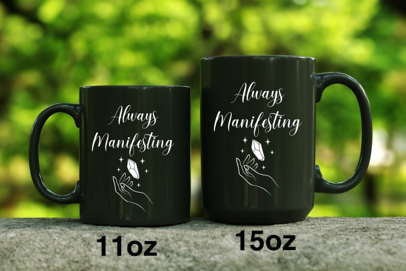 May include: Two black ceramic mugs with white text and a crystal design. The text on the mugs reads "Always Manifesting". The mug on the left is 11 oz and the mug on the right is 15 oz.