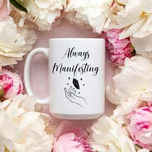 May include: White ceramic mug with a black design that says "Always Manifesting" with a hand holding a crystal and stars. The mug is surrounded by white and pink peonies.