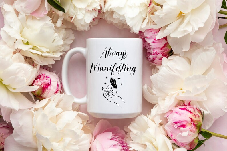 May include: White ceramic mug with a black design that says "Always Manifesting" with a hand holding a crystal and stars. The mug is surrounded by white and pink peonies.