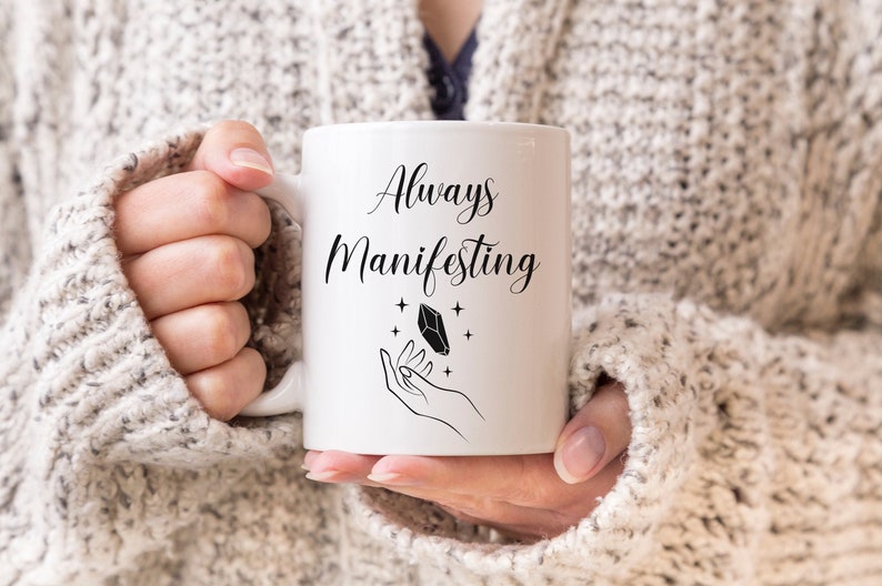 May include: White ceramic mug with black text that reads "Always Manifesting". A black and white illustration of a hand holding a crystal with stars around it is on the mug.