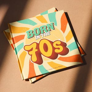 May include: A book with the title "Born in the 70s" in a retro style. The cover has a sunburst design in orange, yellow, and teal, with the title in a 3D font. The book is on a brown surface.
