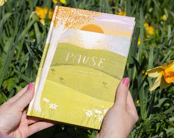 Może przedstawiać: Książka w twardej oprawie zatytułowana PAUSE A WELLBEING WORKBOOK z ilustracją krajobrazu przedstawiającą zielone wzgórze, duże drzewo z pomarańczowymi liśćmi i zachodzące słońce. Okładka zawiera białe kwiaty stokrotki na dole i jasne niebo.