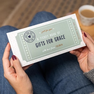 May include: A rectangular gift book with a mint green and white design. The text reads "A Ticket Book of Gifts for Grace" with the name "Yasmin" below. The book is held by a person wearing blue jeans.