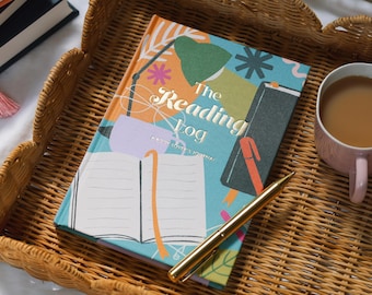 May include: A decorative wicker tray holds a reading log book, a gold pen, and a pink mug of coffee. The book cover is teal with the words "The Reading Log" and colorful illustrations. A stack of books is visible in the background.
