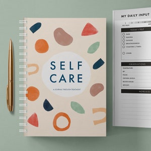 May include: A self-care journal with a beige cover featuring colourful abstract shapes and the text "SELF CARE A JOURNAL THROUGH TREATMENT". The journal has a spiral binding and is open to a page with a daily input section and an observations section.