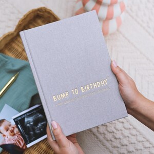May include: A light gray journal titled "Bump to Birthday" with gold lettering. The journal is held open, revealing the cover's title. The journal is a pregnancy and first year journal. A photo of a baby and an ultrasound image are visible.