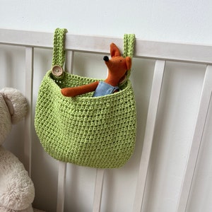 May include: A lime green crochet storage bag with a button closure, hanging on a white crib. Inside the bag is a stuffed fox toy wearing denim overalls. The bag is designed to hold small items.