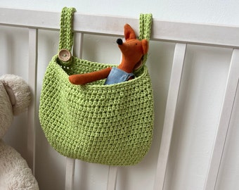 May include: A lime green crochet storage bag with a button closure, hanging on a white crib. Inside the bag is a stuffed fox toy wearing denim overalls. The bag is designed to hold small items.