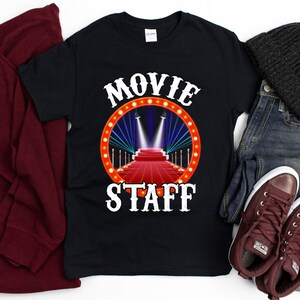 May include: Black t-shirt with white text that reads "MOVIE STAFF" and a graphic of a red carpet with spotlights.