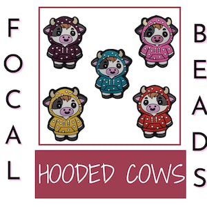 以下が含まれることがあります： 異なる色のフード付きスウェットシャツを着た、漫画の牛のチャームが5つ。チャームは紫、青、黄、ピンク、赤。チャームは下部に「HOODED COWS」と綴られています。