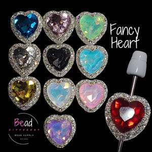 May include: A collection of heart-shaped charms with faceted centers in various colors, each bordered by a silver-toned frame with clear crystals. One red heart charm is attached to a metal pin. The text Fancy Heart and Bead Different Bead Supply Est 2021 are visible.