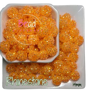 May include: A bowl filled with 14mm orange rhinestone beads. The beads have a sparkly finish and are perfect for crafting.