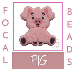 May include: Pink silicone pig bead with a cartoon face and a small pink snout. The bead is sitting on a pink surface with the word "PIG" written in white.