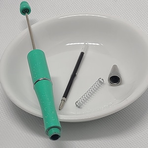 May include: A disassembled turquoise glitter pen with a silver metal tip, a metal spring, and a black ink refill. The pen body features a sparkly finish and a silver center band. The components are laid out separately to show the internal parts.