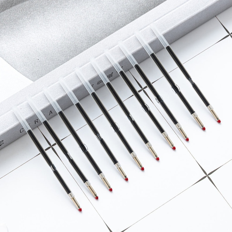 May include: A set of nine black ink pen refills, each featuring a clear plastic protective cap over the tip. The refills have a slender cylindrical body filled with black ink and a metallic tip with a small red wax seal at the very end.
