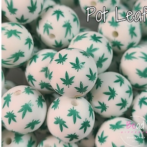 May include: A collection of white, round beads with a green marijuana leaf pattern. The beads have a hole for stringing and the text "Pot Leaf" is visible in the image.