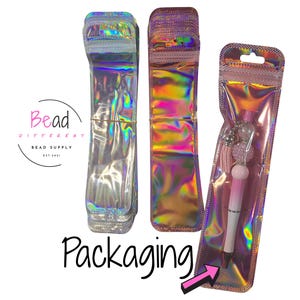 Op de afbeelding: Holografische verpakking voor pennen. De afbeelding toont drie iriserende zakjes, waarvan twee leeg en één met een roze en witte pen met decoratieve kralen. Het woord "Packaging" staat in zwart script onder de zakjes. Het logo van "Bead Different Bead Supply" is ook zichtbaar.