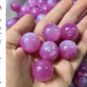 May include: Close-up of several round, purple acrylic beads with a glittery, iridescent effect. The beads are held in a hand, with the word "ACRYLIC" written vertically on the left side of the image. The beads are for jewelry making.