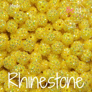 Puede incluir: Primer plano de un montón de cuentas redondas amarillas con un acabado brillante. Las cuentas tienen un diámetro de 14 mm y están etiquetadas como "Rhinestone".