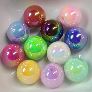Puede incluir: Una colecci&oacute;n de cuentas esf&eacute;ricas iridiscentes en varios colores, incluyendo rosa, morado, verde y amarillo. Las cuentas tienen un acabado brillante y parecen estar hechas de vidrio o un material similar. Est&aacute;n dispuestas en un cuenco blanco.