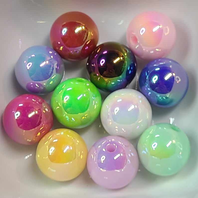 Puede incluir: Una colecci&oacute;n de cuentas esf&eacute;ricas iridiscentes en varios colores, incluyendo rosa, morado, verde y amarillo. Las cuentas tienen un acabado brillante y parecen estar hechas de vidrio o un material similar. Est&aacute;n dispuestas en un cuenco blanco.