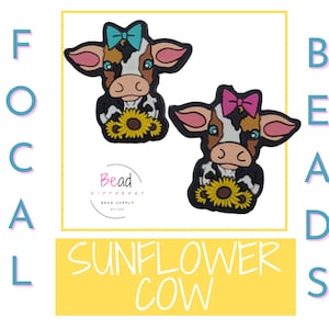 以下が含まれることがあります： 口にひまわりを持った黒と白の牛のチャームが2つ。牛はリボンをつけていて、1つは青、もう1つはピンク。チャームには「Sunflower Cow」と「Bead Divas」のラベルが貼られています。