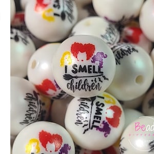 May include: White acrylic beads with a Halloween theme. Each bead features the phrase "I SMELL children" with illustrations of three witches in red, yellow, and purple. The word "Acrylic" is written vertically on the left.