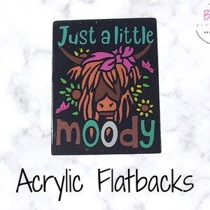 Pode incluir: Base plana de acrílico preto com um desenho de vaca Highland de desenho animado. A vaca tem pelo castanho, um laço rosa e o texto "Just a little moody". Flores decorativas e texto são em verde-azulado, rosa e castanho.