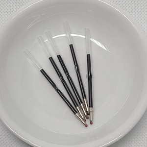 May include: Five clear plastic pen ink refills with black ink reservoirs and metallic tips. Each refill features a transparent tube section and a central crimped area, designed for standard ballpoint pens, arranged in a cluster.