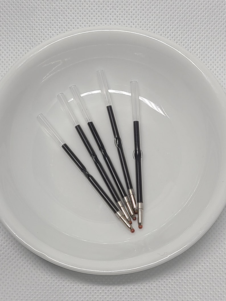May include: Five clear plastic pen ink refills with black ink reservoirs and metallic tips. Each refill features a transparent tube section and a central crimped area, designed for standard ballpoint pens, arranged in a cluster.