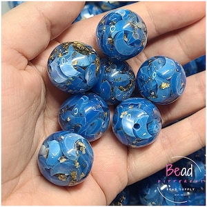 May include: A close-up of blue acrylic beads with gold flecks. The beads are round and have a marbled appearance. The beads are being held in a hand.