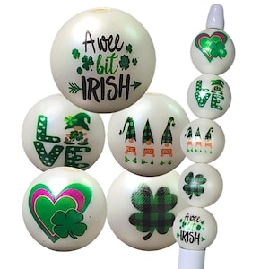 May include: A collection of white, round beads with various St. Patrick's Day designs. The beads feature shamrocks, gnomes, hearts, and the text "A wee bit Irish" and "Love". The beads are for crafting or decorative purposes.