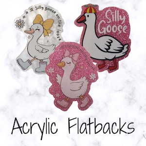 Può includere: Tre decorazioni piatte in acrilico con oche dei cartoni animati e la scritta "Silly Goose on the Loose". Un'oca indossa un fiocco e stivali gialli, un'altra un cappello rosso e giallo, e la terza un fiocco e stivali rosa. Su sfondo marmoreo.