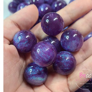 May include: Close-up of several purple acrylic beads with a shimmering, iridescent effect. The beads are spherical and held in a hand. The word "Acrylic" is written vertically on the left side of the image. The Bead Supply logo is in the bottom right corner.