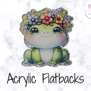 May include: Acrylic flatback featuring a cute frog with a floral crown. The frog is green with blue eyes and pink cheeks, surrounded by a silver glitter border. The text "Acrylic Flatbacks" is at the bottom.
