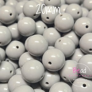 Puede incluir: Un montón de cuentas redondas grises claras de 20 mm con un agujero central. Las cuentas son lisas y brillantes. El texto "20mm" está en blanco en la parte superior de la imagen. El texto "Bead Different" y "Bead Supply" está en la esquina inferior derecha.