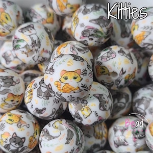 Op de afbeelding: Witte, ronde kralen met kattenillustraties in verschillende kleuren. De kralen hebben een glanzende afwerking. Het woord "Kitties" staat in cursief bovenaan. Deze kralen zijn voor het maken van sieraden.