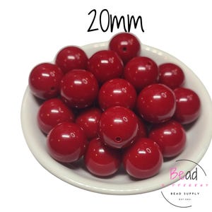 Puede incluir: Un cuenco blanco lleno de cuentas redondas rojas brillantes de 20 mm. Las cuentas tienen una superficie lisa y pulida con un agujero en el centro. El texto "20mm" está encima de las cuentas. La esquina inferior derecha tiene el texto "Bead Different Bead Supply EST 2021".