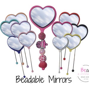 以下が含まれることがあります： ピンク、青、黄色など、さまざまな色のハート型のハンドミラーのコレクション。各ミラーには細長い柄があり、ベースには装飾的なビーズが付いています。「Beadable Mirrors」というテキストが下に表示されています。